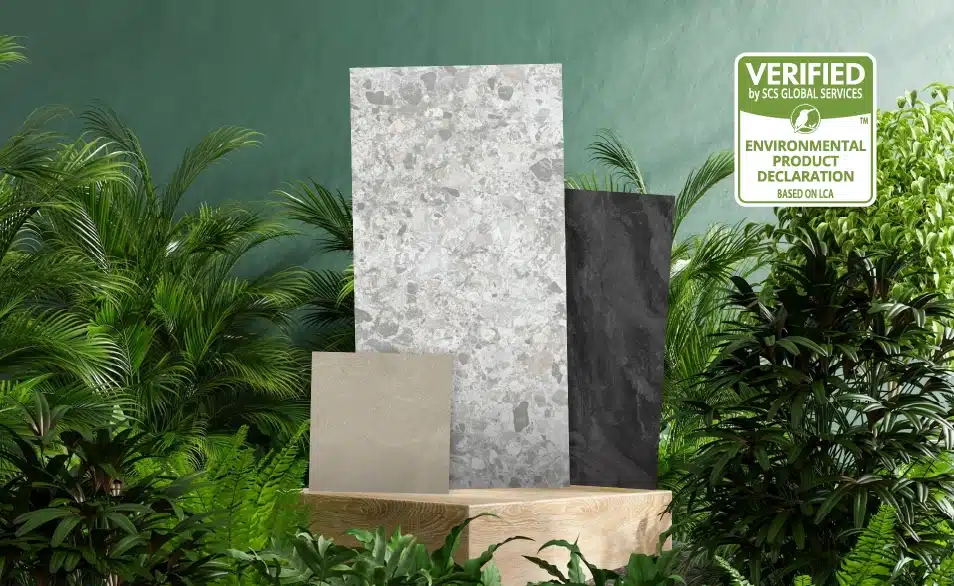 A small green-coloured labeling that denotes 'Environmental Product Declaration' and is verified by SCS Global Services is seen, where three different design types and sizes of Niro Granite's tiles are placed on top of a wooden block that is surrounded by a green bush of leaves. This shows that Niro Granite's tiles are eco-friendly in manufacturing.