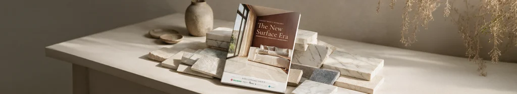 Wide website banner featuring 'The New Surface Era' brochure leaning against stacked natural stone and marble tiles on a neutral, minimalist desk.